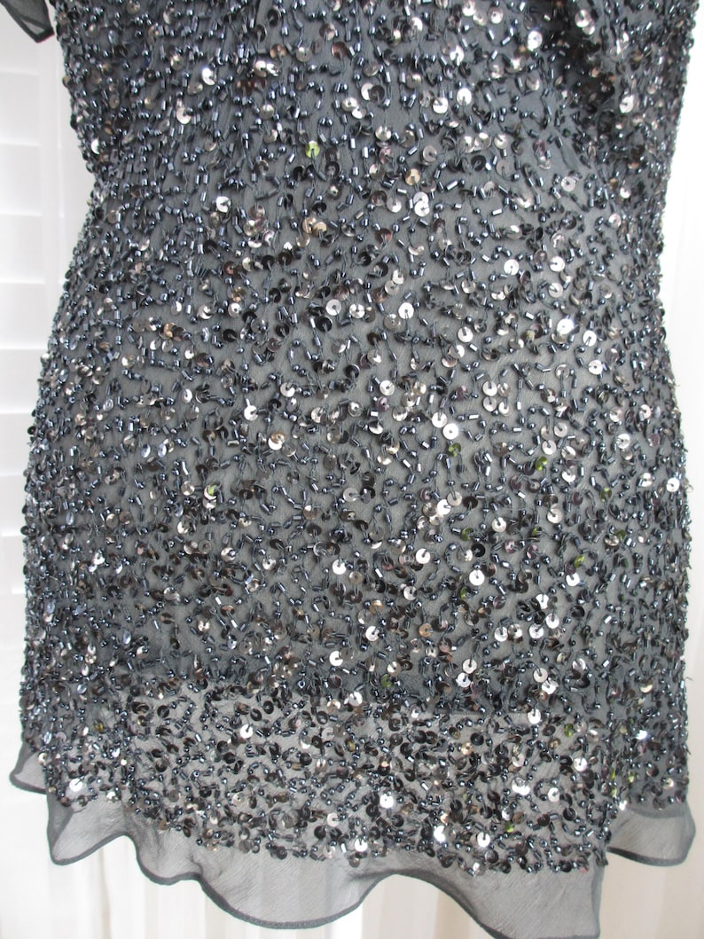 Puede incluir: Top gris oscuro, ricamente adornado con lentejuelas y abalorios. La prenda tiene un dobladillo transparente con volantes y est&aacute; cubierta de peque&ntilde;os adornos reflectantes. El top parece estar hecho de una tela ligera.
