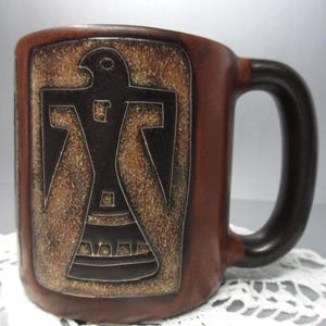 Peut inclure: Mug en céramique marron avec une anse marron foncé. Le mug présente un motif rectangulaire avec un emblème d'oiseau noir stylisé. Le fond a un aspect texturé et vieilli. Le mug est posé sur un napperon en dentelle blanche.