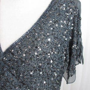 Puede incluir: Un vestido gris oscuro con cuentas, con escote en V y mangas cortas. El vestido est&aacute; cubierto de peque&ntilde;as cuentas y lentejuelas brillantes, creando un efecto texturizado y brillante. El escote est&aacute; adornado con cuentas m&aacute;s oscuras.