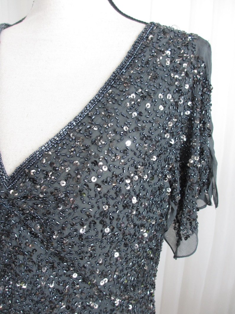 Puede incluir: Un vestido gris oscuro con cuentas, con escote en V y mangas cortas. El vestido est&aacute; cubierto de peque&ntilde;as cuentas y lentejuelas brillantes, creando un efecto texturizado y brillante. El escote est&aacute; adornado con cuentas m&aacute;s oscuras.