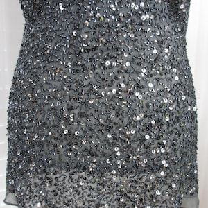 Puede incluir: Un vestido gris oscuro cubierto de lentejuelas y abalorios. El vestido tiene un escote redondo y mangas cortas. La tela es un material transparente y ligero. El dobladillo es festoneado.