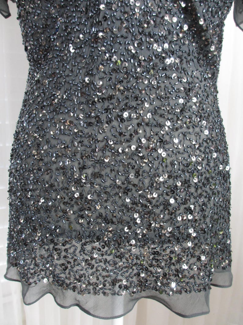 Puede incluir: Un vestido gris oscuro cubierto de lentejuelas y abalorios. El vestido tiene un escote redondo y mangas cortas. La tela es un material transparente y ligero. El dobladillo es festoneado.