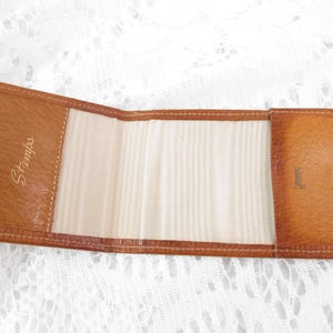 Puede incluir: Un port sellos de cuero marrón vintage, abierto y plano. El interior es de color beige claro con un patrón texturizado. La palabra "Stamps" está grabada en dorado en el lado izquierdo. Se ve un cierre de botón a presión dorado a la derecha.