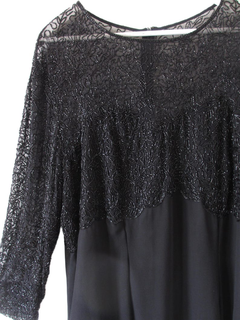 Puede incluir: Vestido negro con mangas largas. La parte superior del vestido presenta intrincados detalles de cuentas, creando un patr&oacute;n texturizado en forma de flor. Las mangas son transparentes y el vestido tiene un cuello redondo.