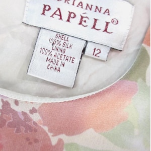 May include: Close-up of a dress tag with the brand name "Adrianna Papell" in red lettering. The tag indicates the shell is 100% silk and the lining is 100% acetate. The size is 12. The dress features a floral pattern in shades of pink and green.
