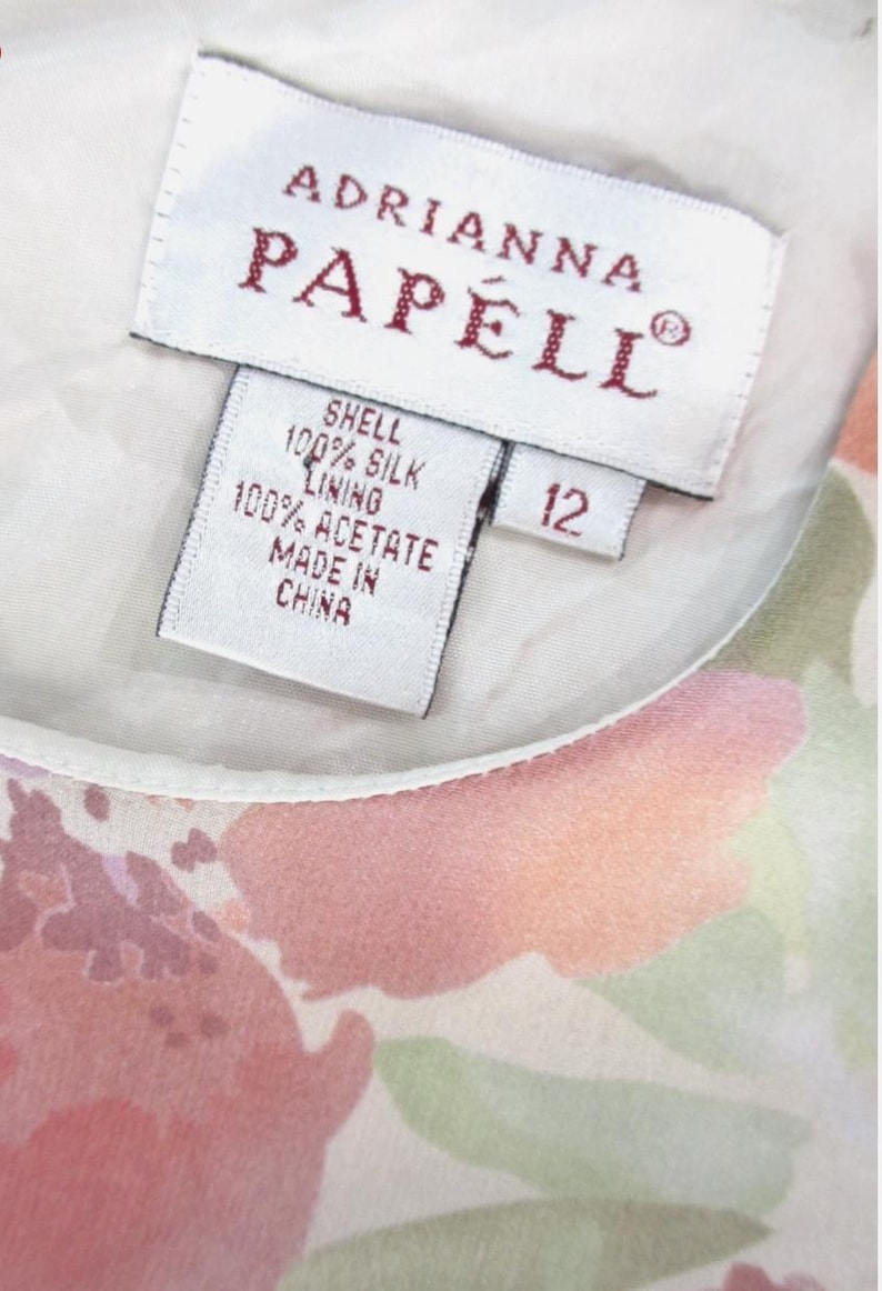May include: Close-up of a dress tag with the brand name "Adrianna Papell" in red lettering. The tag indicates the shell is 100% silk and the lining is 100% acetate. The size is 12. The dress features a floral pattern in shades of pink and green.