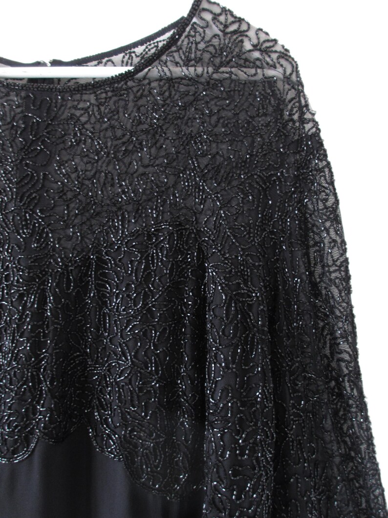 Puede incluir: Top de noche negro con intrincados patrones florales de cuentas. El top presenta un escote y mangas transparentes, con un dobladillo festoneado. El abalorio a&ntilde;ade textura y brillo a la prenda, creando un look elegante y sofisticado.