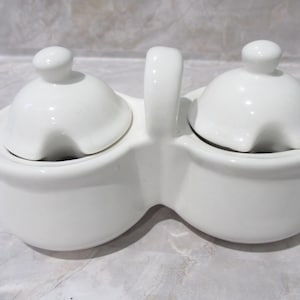 Puede incluir: Un juego de salero y pimentero de cerámica blanca. El juego presenta dos compartimentos redondeados con tapas abovedadas a juego y un asa central. Las tapas tienen una pequeña muesca para el acceso de la cuchara.