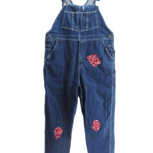 May include: Blue denim overalls with adjustable straps, multiple pockets, and red bandana patches on the knees and upper thigh. The overalls have a classic workwear style with metal hardware and a relaxed fit.