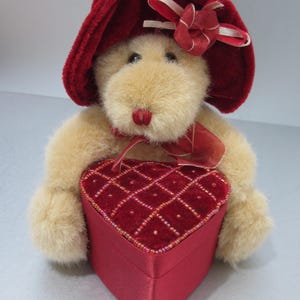 May include: A plush teddy bear wearing a red hat and holding a heart-shaped box. The box is red with a velvet top and beaded accents. The bear has a red nose and bow.