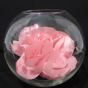 May include: A clear, round glass bowl with a large pink flower inside. The flower has multiple petals and is the focal point of the image. The bowl sits on a black surface.