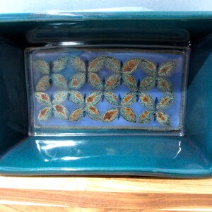 May include: Teal ceramic rectangular baking dish with a light blue interior. The interior has a decorative pattern of brown and teal leaf-like shapes. The dish has handles and is on a wooden surface.