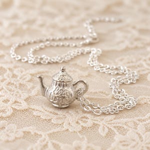 May include: A silver-toned metal necklace featuring a miniature teapot pendant. The teapot has a curved spout, a handle, and a lid, with the words "tea time" embossed in script lettering on the front surface. The pendant hangs from a delicate silver chain.