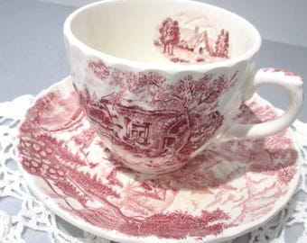 Può includere: Un set di tazza e piattino bianchi con un disegno rosso transferware raffigurante un paesaggio rurale. La tazza ha un manico e poggia su un piattino abbinato. Il set è posto su una tovaglietta di pizzo bianco.
