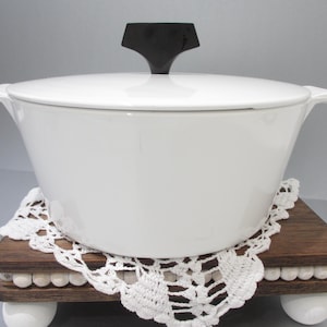May include: A white enamel Dutch oven with a black handle, resting on a wooden trivet with a white lace doily. The trivet has white ball feet and is made of dark wood.