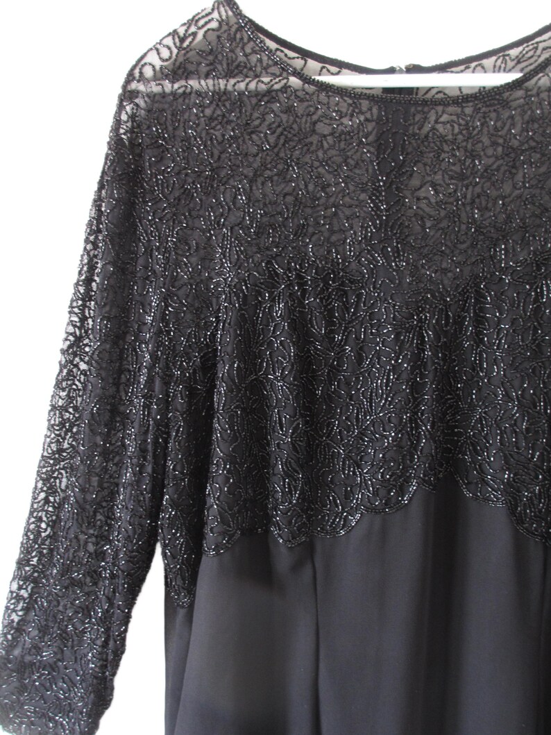 Puede incluir: Vestido de noche negro con mangas largas. La parte superior del vestido presenta intrincados detalles de cuentas, creando un efecto texturizado y brillante. Las mangas son transparentes y la falda es opaca.