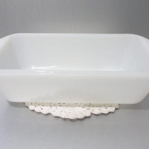 May include: A rectangular, white, vintage baking dish with rounded corners. The dish is resting on a white, crocheted doily. The dish is made of opaque glass and has a smooth, glossy finish. The overall aesthetic is simple and classic.