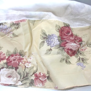 May include: A floral fabric with a cream-colored background and a pattern of large, red and pink roses, purple hydrangeas, and green leaves. The fabric is draped, with a white fabric visible at the top.