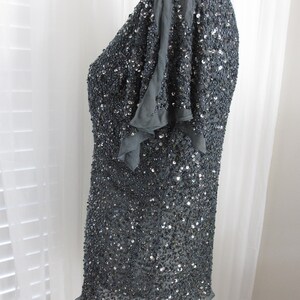 Puede incluir: Una blusa gris oscuro, muy adornada con lentejuelas, con mangas cortas y con volantes. La prenda est&aacute; cubierta de peque&ntilde;as lentejuelas reflectantes, creando un efecto brillante. El dobladillo tiene un borde delicado y con volantes. La blusa se muestra en un maniqu&iacute;.