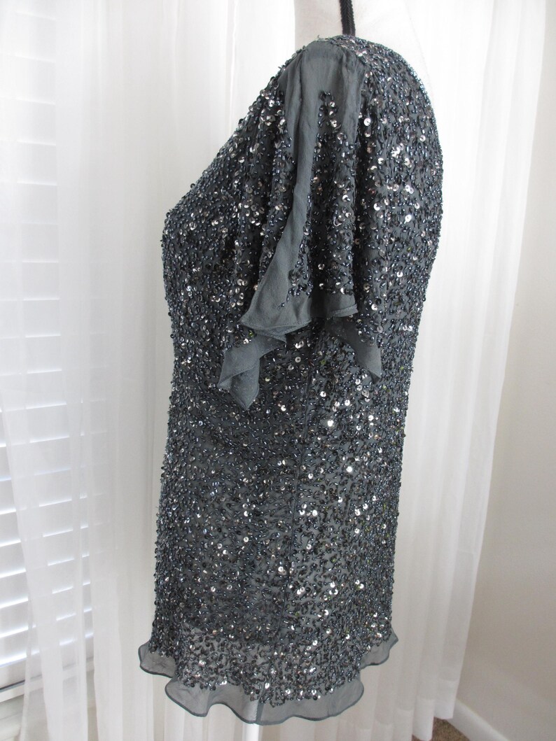 Puede incluir: Una blusa gris oscuro, muy adornada con lentejuelas, con mangas cortas y con volantes. La prenda est&aacute; cubierta de peque&ntilde;as lentejuelas reflectantes, creando un efecto brillante. El dobladillo tiene un borde delicado y con volantes. La blusa se muestra en un maniqu&iacute;.