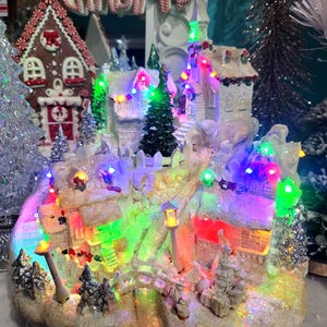 May include: A Christmas village scene with illuminated houses, trees, and a bridge. The miniature houses have snow-covered roofs and are decorated with colorful lights. The scene includes a gingerbread house and a sign that says "MERRY CHRISTMAS".