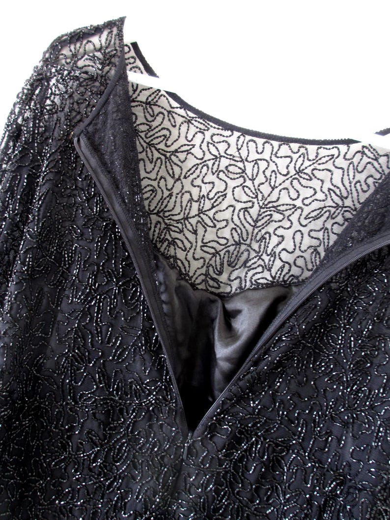Puede incluir: Primer plano de un vestido negro con cuentas y un detallado estampado floral. El vestido presenta un escote en V profundo y cierre de cremallera. El forro es visible, mostrando una tela blanca contrastante con un dise&ntilde;o de hojas negras.