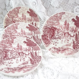May include: Set of four white ceramic plates with a red transferware design. Each plate features a detailed pastoral scene with trees, buildings, and figures. The plates are arranged on a white lace surface.