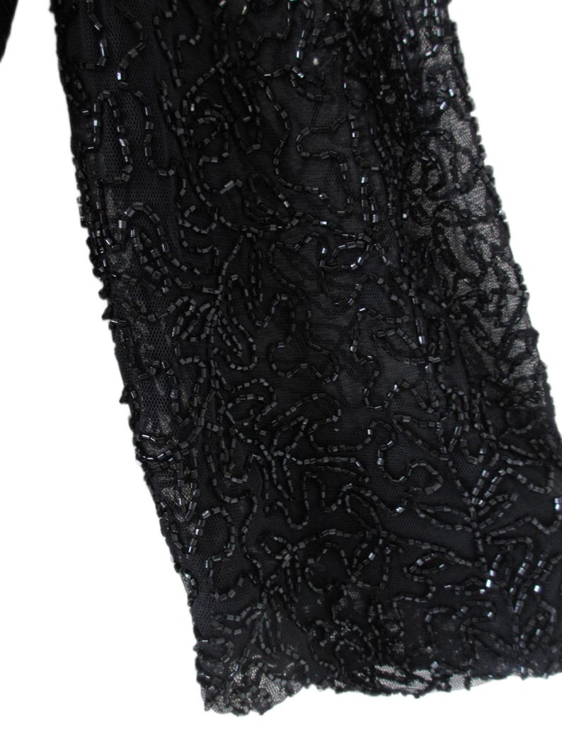 Puede incluir: Primer plano de una manga negra, ricamente adornada con intrincados abalorios. Las cuentas est&aacute;n dispuestas en un patr&oacute;n arremolinado, similar a flores, creando un dise&ntilde;o texturizado y elegante. La manga est&aacute; hecha de una tela negra transparente.