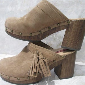 May include: A pair of tan suede clogs with wooden heels. The clogs have a rounded toe, a strap across the top, and are adorned with small metal studs. One clog features decorative tassels. The heels are approximately 4 inches high.