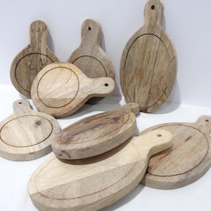 Può includere: Una collezione di sette taglieri da portata in legno marrone chiaro con manici. Ogni tagliere presenta un'area incassata circolare o ovale sulla superficie e un piccolo foro nella parte superiore del manico per appenderlo. La venatura è visibile.