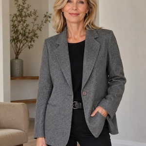 May include: A charcoal grey wool-blend blazer featuring a notched lapel, a single-button front closure, and two side pockets. The blazer is worn over a black top and black trousers with a black leather belt, creating a structured and layered look.