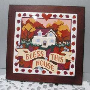 May include: A square, framed tile with a cross-stitch design. The image features a white house with a purple roof, surrounded by autumn trees. The words "BLESS THIS HOUSE" are displayed on a banner, framed by leaves and flowers. The frame is dark brown.