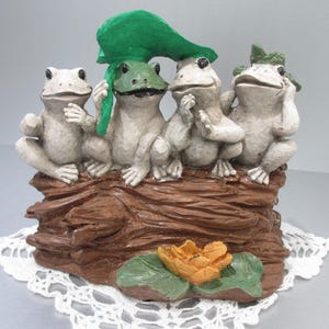 May include: A whimsical figurine featuring four frog figures perched on a brown, textured log. One frog holds a green leaf overhead. A small orange flower and green leaves are at the base. The figurine sits on a white lace doily.