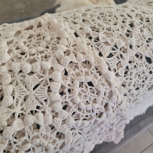 May include: An off-white crochet blanket featuring an intricate, repeating floral pattern with raised bobble stitch details. The textile is crafted with a complex openwork design, showcasing a dense arrangement of interconnected circular motifs.