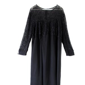Puede incluir: Vestido negro con mangas largas. La parte superior del vestido presenta un dise&ntilde;o texturizado y de encaje, mientras que la parte inferior es de tela lisa y s&oacute;lida. El vestido cuelga de una percha blanca sobre un fondo blanco.