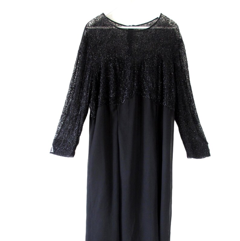 Puede incluir: Vestido negro con mangas largas. La parte superior del vestido presenta un dise&ntilde;o texturizado y de encaje, mientras que la parte inferior es de tela lisa y s&oacute;lida. El vestido cuelga de una percha blanca sobre un fondo blanco.