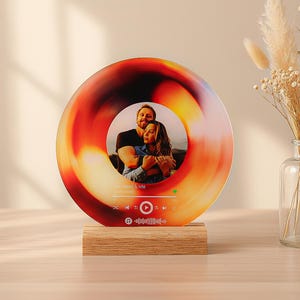 May include: A round, colorful photo frame with a picture of a couple. The frame has a gradient of orange, red, and yellow. It sits on a wooden base. The frame has a music player interface.