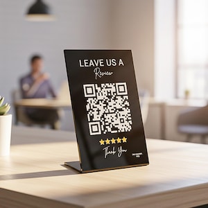 May include: A black table tent with the text "LEAVE US A Review" and "Thank You" in white. A QR code is in the center, with five gold stars below. The sign is on a light-colored wooden table.