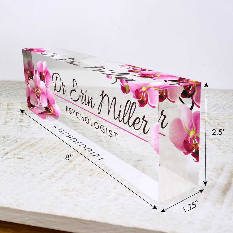 May include: A clear acrylic desk nameplate with a pink orchid floral design. The nameplate reads "Dr. Erin Miller, Psychologist". The nameplate measures 8 inches long, 2.5 inches tall, and 1.25 inches wide.