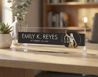 May include: A clear acrylic desk nameplate with the name "EMILY K. REYES" and the title "ATTORNEY AT LAW" in white text on a black background. A gold-colored emblem of a person holding scales is on the right side.