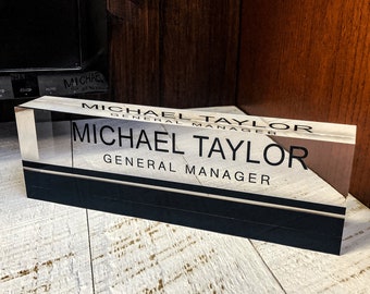Op de afbeelding: Een helder acryl naamplaatje voor op het bureau met zwarte letters. Het naamplaatje heeft de tekst "Michael Taylor, General Manager".