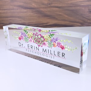 May include: A clear acrylic desk nameplate with a floral design featuring succulents in shades of pink, green, and purple. The text "Dr. Erin Miller, Psychologist" is printed in black on the nameplate.