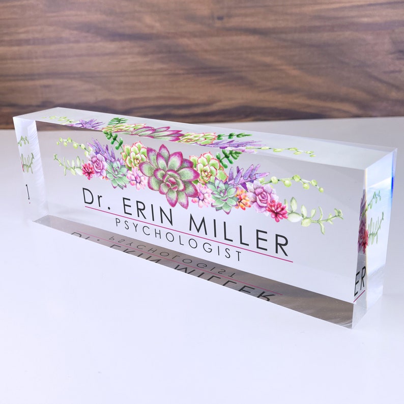 May include: A clear acrylic desk nameplate with a floral design featuring succulents in shades of pink, green, and purple. The text "Dr. Erin Miller, Psychologist" is printed in black on the nameplate.
