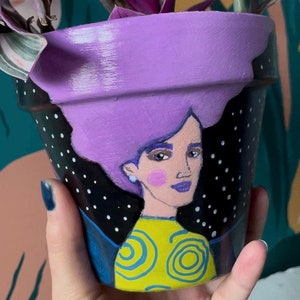 Puede incluir: Maceta de terracota pintada de morado con fondo de lunares negros y blancos. La maceta está pintada con un retrato de una mujer con cabello morado, una camisa amarilla con remolinos azules y rubor rosado.