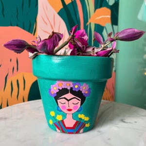 Puede incluir: Maceta de terracota pintada en turquesa con un retrato pintado a mano de Frida Kahlo con una corona de flores. La maceta tiene flores amarillas pintadas en los lados.