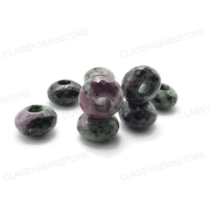 May include: A collection of faceted gemstone beads. The beads are a mix of colors, including deep red, green, and black. Each bead has a hole running through the center, and they are scattered on a white surface.