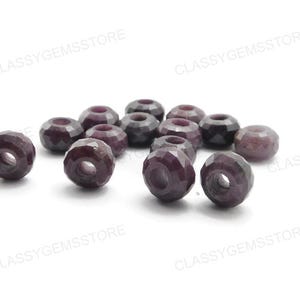 Puede incluir: Una colección de cuentas facetadas de piedras preciosas de color púrpura oscuro con un agujero en el centro. Las cuentas están dispersas sobre una superficie blanca. Las cuentas tienen aproximadamente 0,8 cm de diámetro.