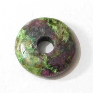 May include: A polished, donut-shaped gemstone with a central hole. The stone features a mix of green, purple, and reddish-brown hues, creating a marbled effect. The overall shape is circular, and the surface appears smooth and glossy.