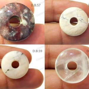Pode incluir: Quatro contas de pedras preciosas em forma de donut. As contas são de várias cores e padrões, incluindo uma pedra manchada marrom-avermelhada, uma pedra branca com veios cinzentos, uma pedra branca com detalhes verdes e uma pedra transparente. Cada conta tem um furo central.