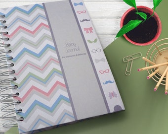 May include: A gray and white chevron patterned baby journal with a gray spine. The spine has a silver spiral binding and features the text "Baby Journal" and "For Cameron & Delaney" in black text. The spine is decorated with small colorful icons, including a bow, glasses, a mustache, and a butterfly.